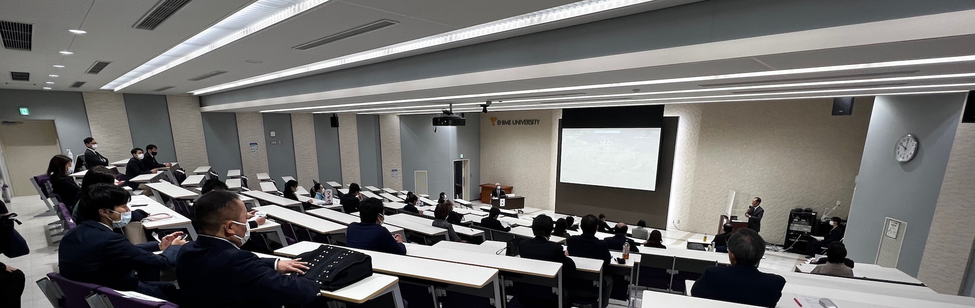 愛媛大学医学部40周年記念講堂の客席後方から演台を見た写真。階段状の座席に参加者が着席し、正面のスクリーンに映像が投影されている。